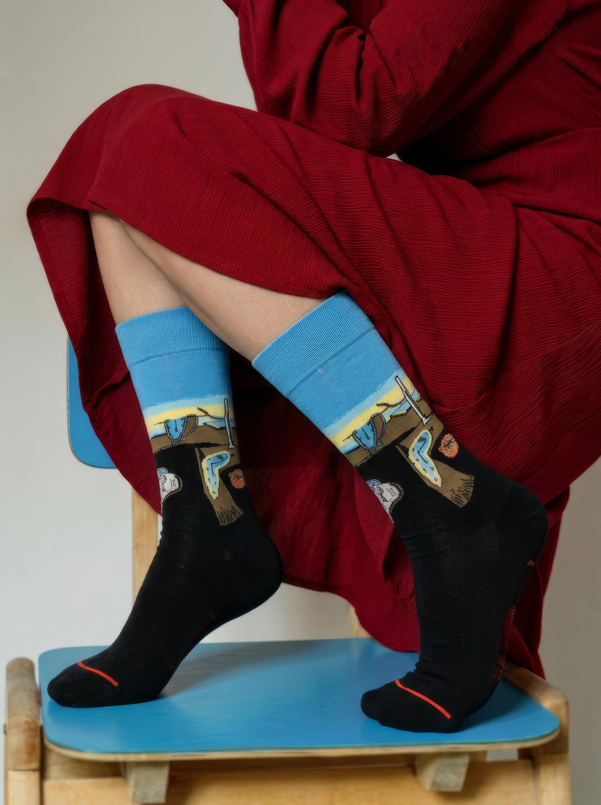 Person in a red dress wearing black XL socks with blue cuffs featuring Salvador Dalí’s surrealist artwork, seated on a blue stool.⠋