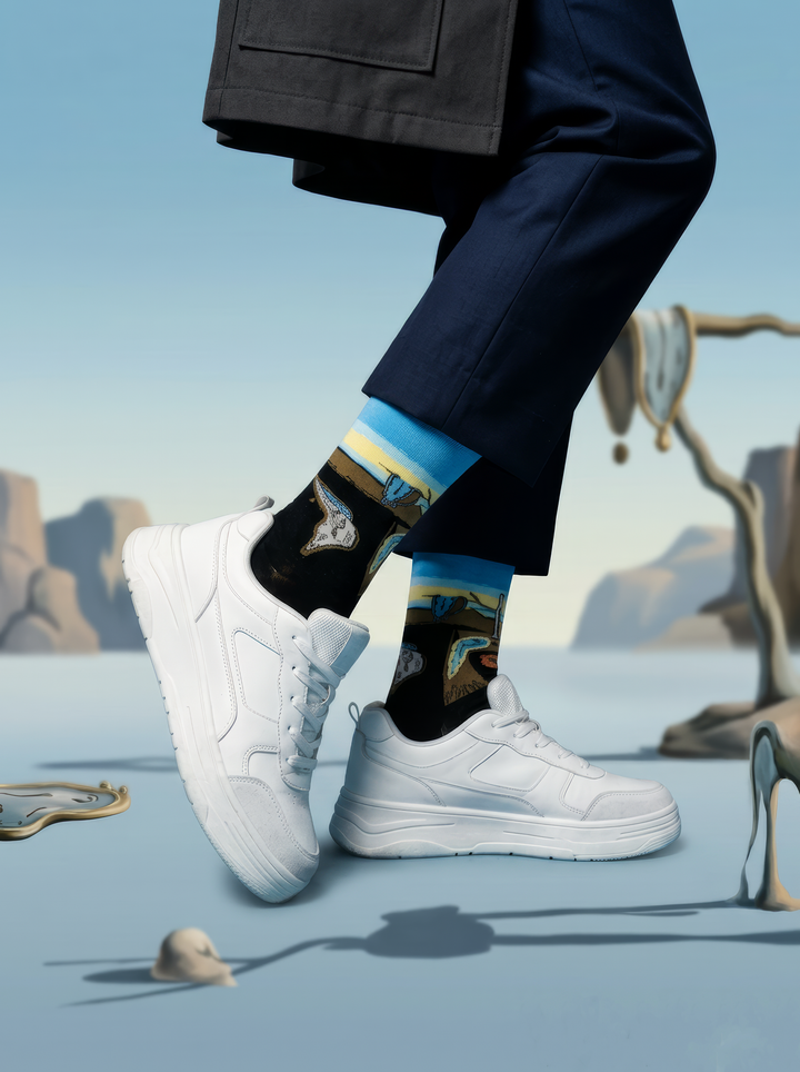 Person in dark trousers wearing black XL socks with blue cuffs and Salvador Dalí artwork, paired with white sneakers against a surreal, melting-clock backdrop.