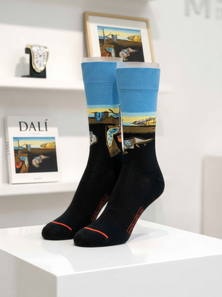Pair of black XL crew socks with blue cuffs featuring colourful Salvador Dalí artwork, displayed on a white pedestal with Dalí books and art objects in the background.