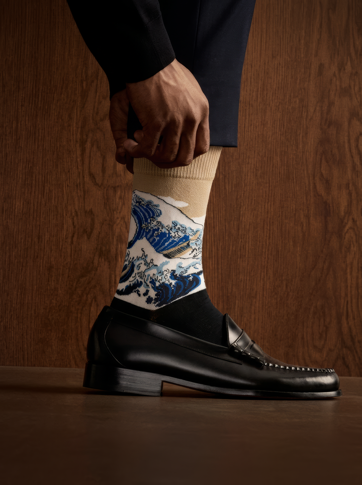 The Great Wave (Classic) art sock by Katsushika Hokusai worn with a black leather shoe against a dark wooden background