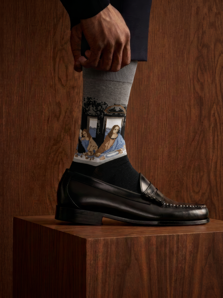The Last Supper art socks featuring Leonardo da Vinci’s artwork worn with a black leather loafer against a wooden background

