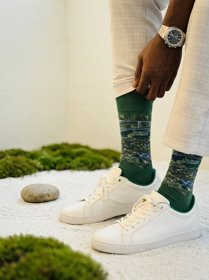 Close-up of green Claude Monet The Japanese Bridge art socks worn with white sneakers beside moss and garden stones. Unisex socks for women and men.
