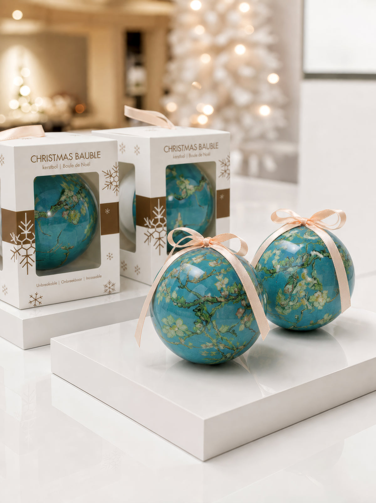Front and back of a Vincent van Gogh Almond Blossom Christmas bauble with cream ribbon on white pedestals with their gift boxes and a white Christmas tree in the background – officially licensed museum art ornament, 7 cm diameter, unbreakable. A unique Dutch Post-Impressionist art Christmas gift from ArtyGifts.


