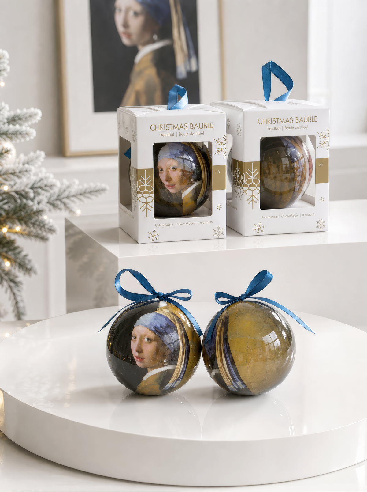 Front and back of a Johannes Vermeer Girl with a Pearl Earring Christmas bauble with blue ribbons displayed on a white pedestal with their gift boxes and the original painting in the background – officially licensed museum art ornament, 7 cm diameter, unbreakable. A unique Dutch Golden Age art Christmas gift from ArtyGifts.


