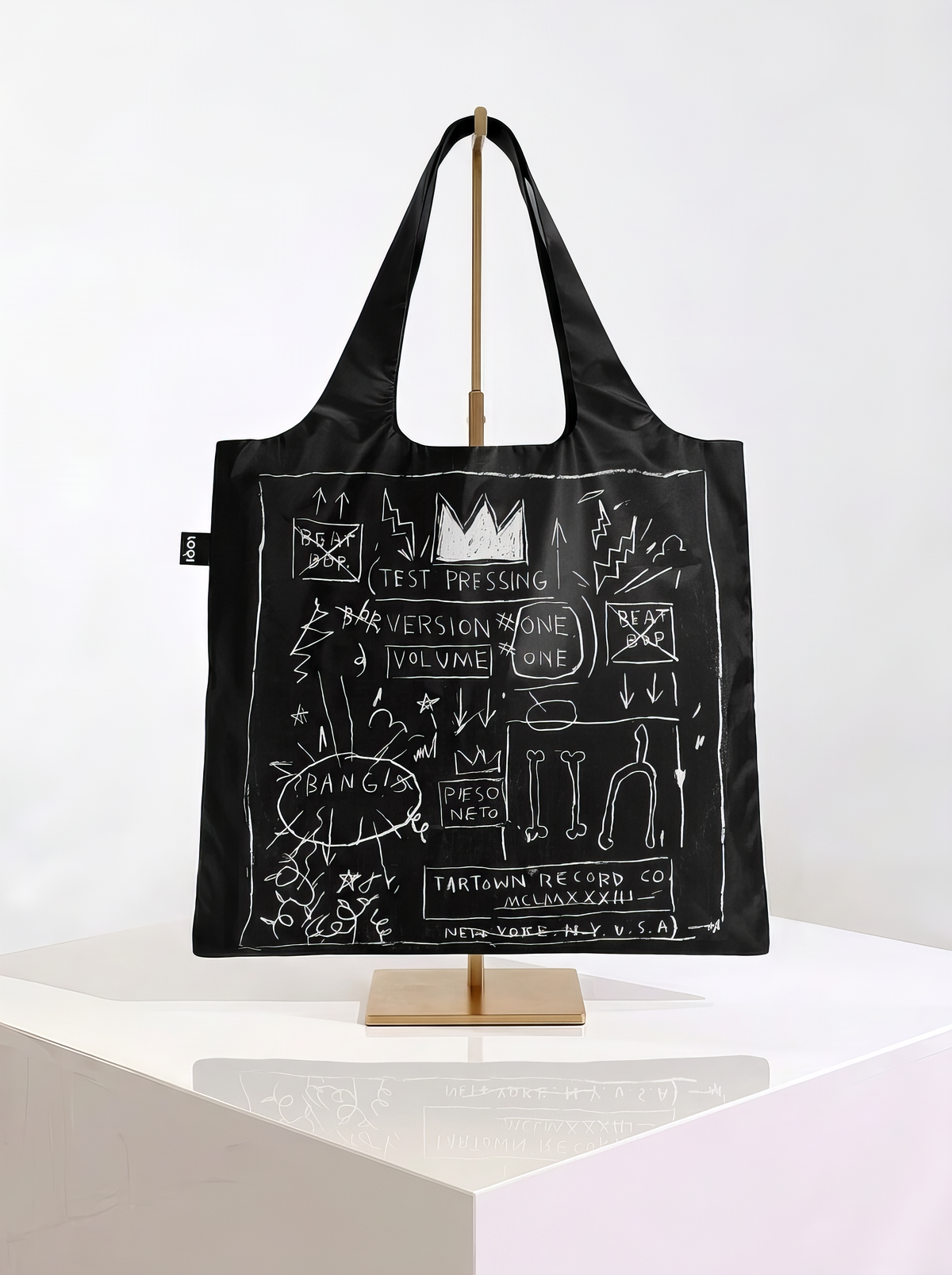 Black LOQI shopper with white crowns, handwritten lettering and line drawings, displayed on a brass-coloured stand on a white pedestal.