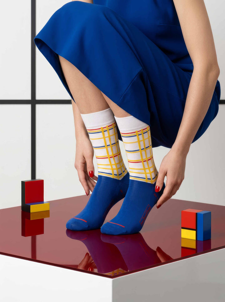 Woman in blue dress wearing MuseARTa Mondrian New York City art socks with Mondrian art background — De Stijl museum gift ArtyGifts
