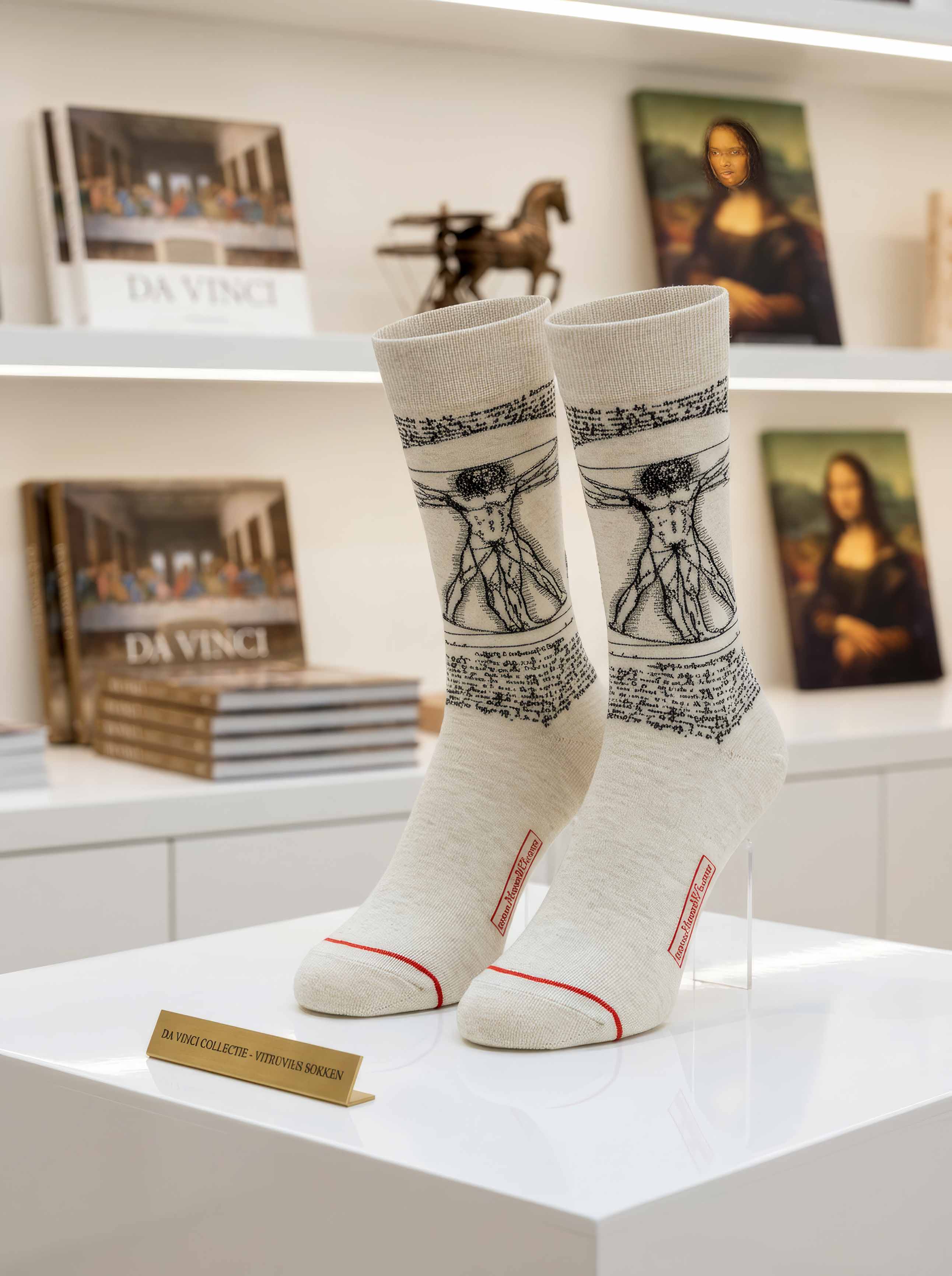 Da Vinci Vitruvian Man art socks on display stand in museum setting with Mona Lisa and Da Vinci artworks in background — ArtyGifts